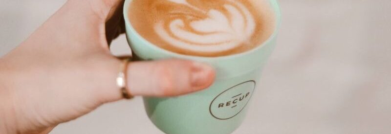 Hand holding a latte with art in a mint reusable cup at a Berlin café. Stylish minimalist scene.