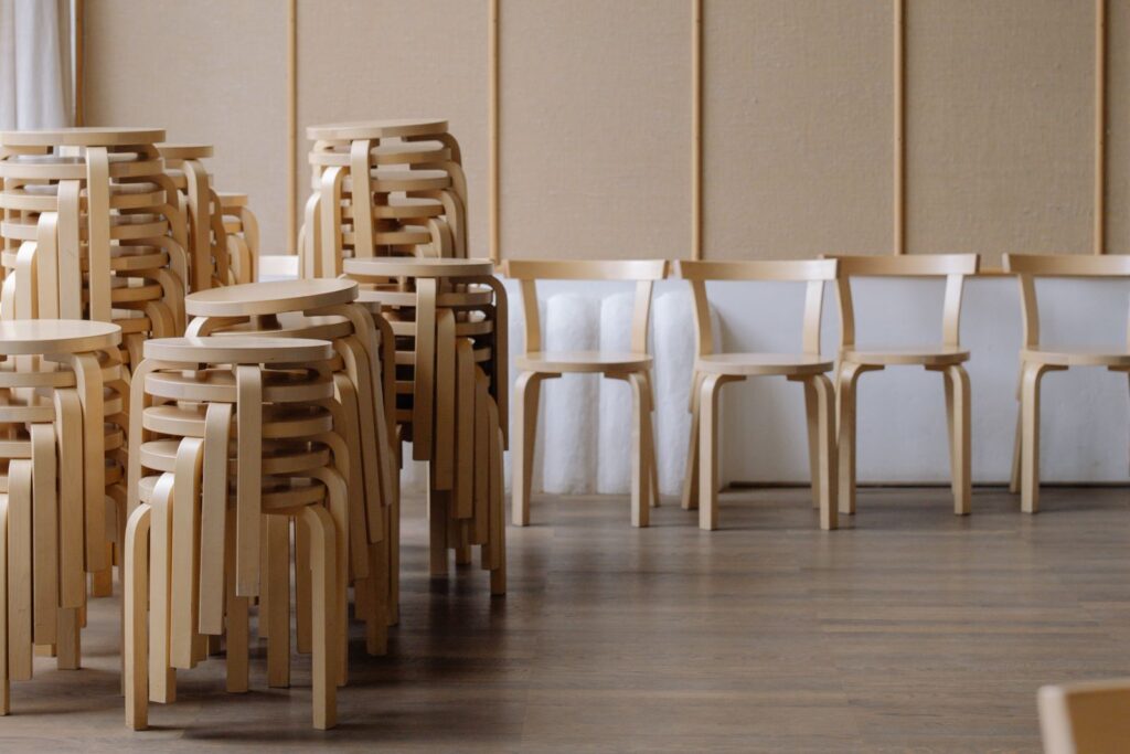 Light-filled minimalist room featuring stacked wooden stools and rows of chairs.